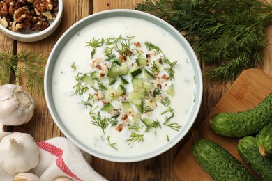 Photo of Tasty cucumber soup with dill and nuts in bowl among products on wooden table, flat lay
