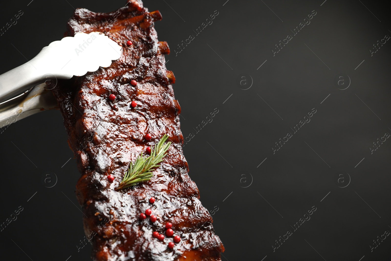 Holding delicious roasted pork ribs with tongs on black background, closeup. Space for text Photo of Holding delicious roasted pork ribs with tongs on black background, closeup. Space for text