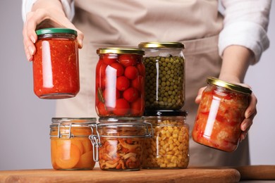 Photo of Woman with jars of different pickled products at table against light grey background, closeup