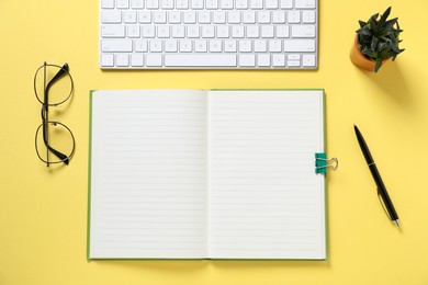Photo of Open blank notebook, computer keyboard, glasses, pen and floral decor on yellow background, flat lay