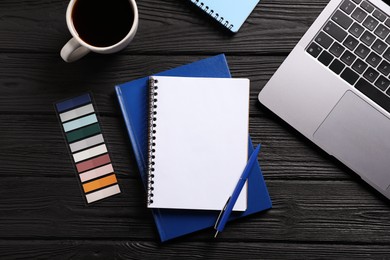 Photo of Open blank notebook, other stationery, coffee and laptop on black wooden table, flat lay