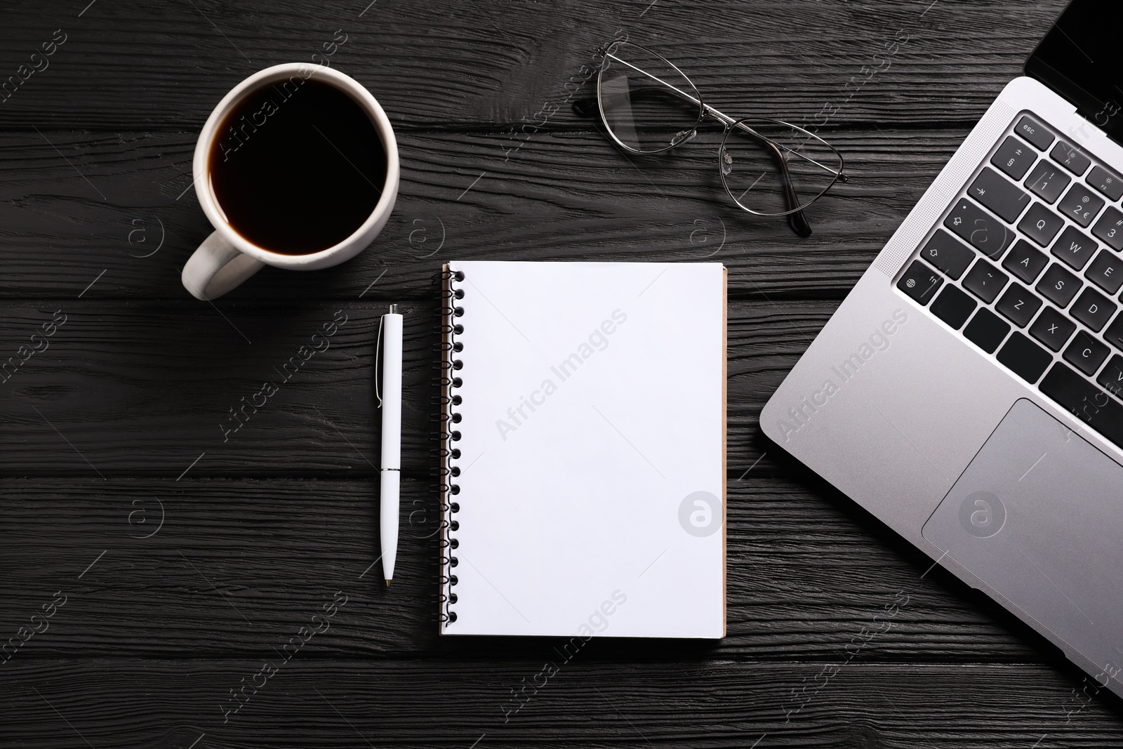 Photo of Open blank notebook, pen, coffee, laptop and glasses on black wooden table, flat lay