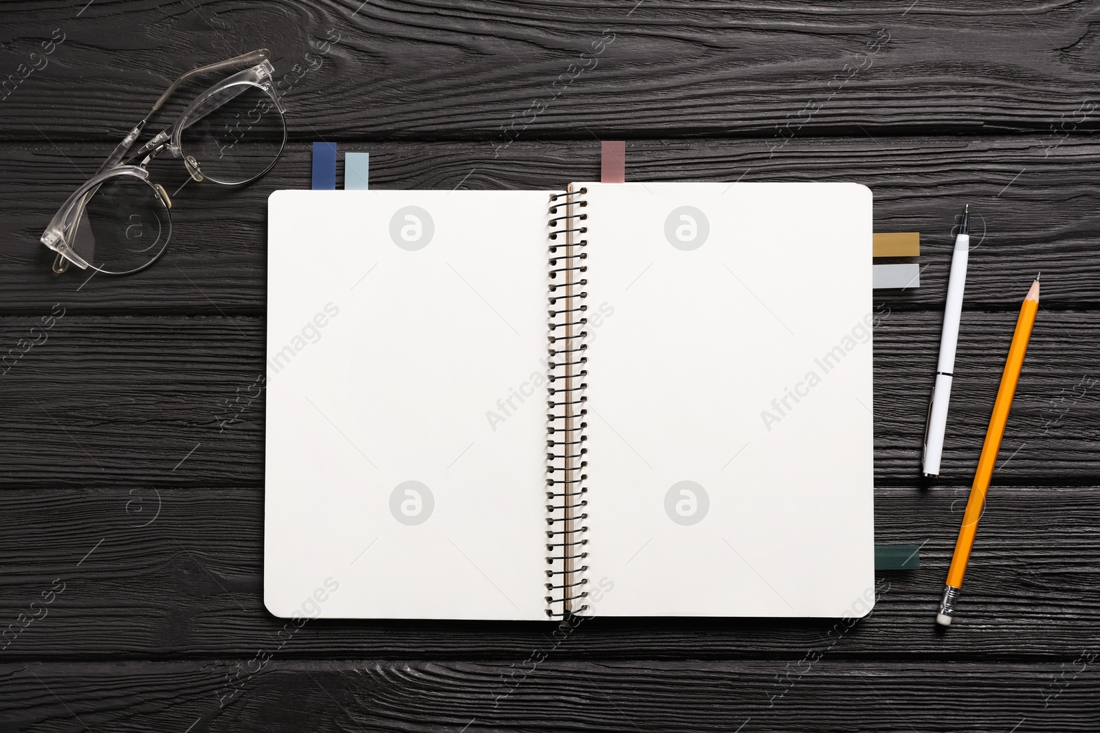 Open blank notebook with colorful stickers, pen, pencil and glasses on black wooden table, flat lay Photo of Open blank notebook with colorful stickers, pen, pencil and glasses on black wooden table, flat lay