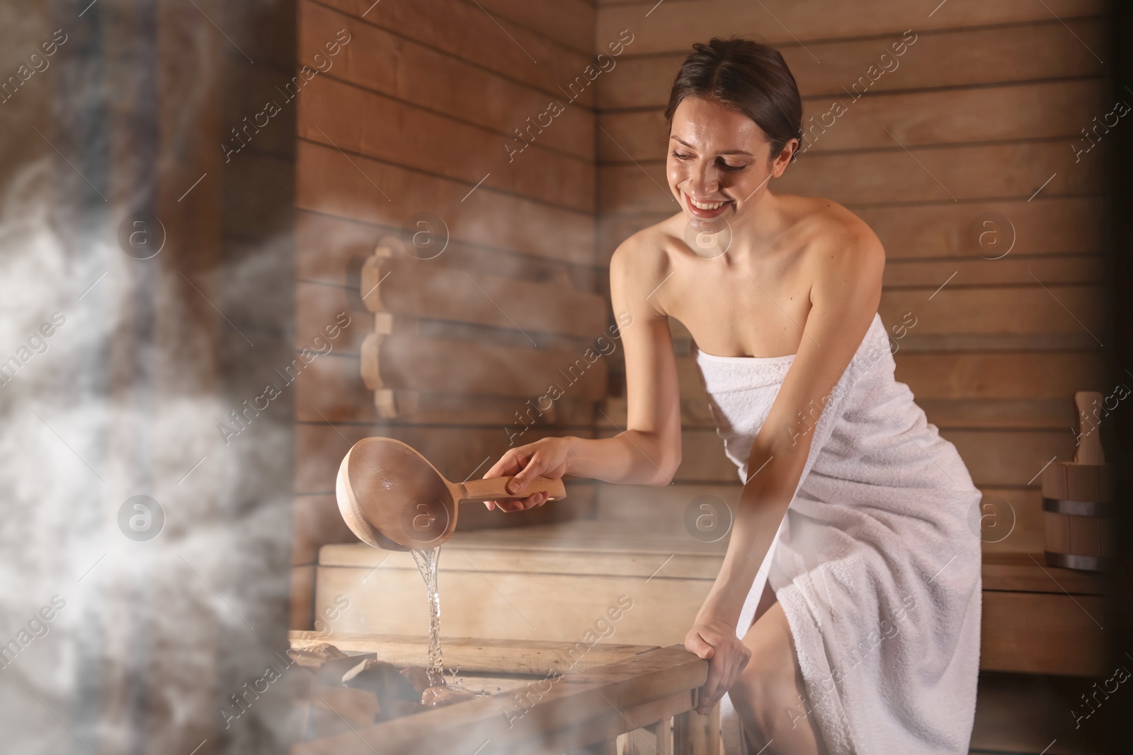 Smiling woman pouring water onto stones surrounded by steam in sauna Image of Smiling woman pouring water onto stones surrounded by steam in sauna