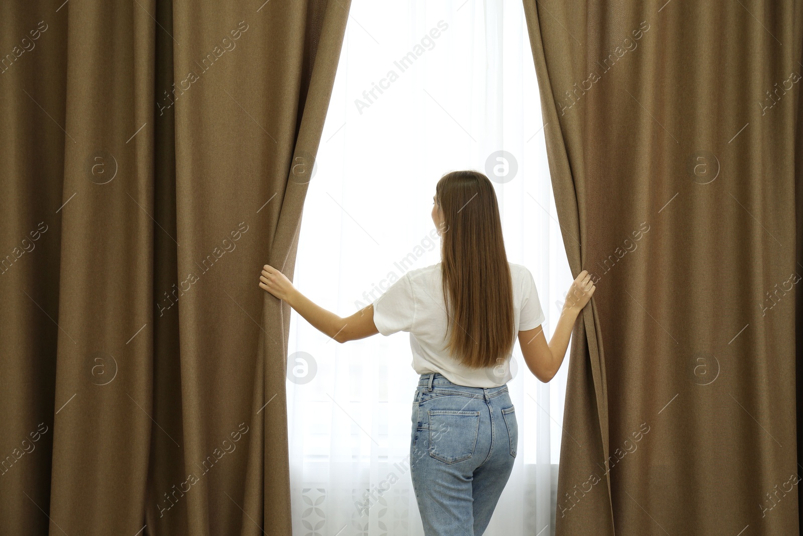 Woman opening elegant window curtains in room, back view Photo of Woman opening elegant window curtains in room, back view