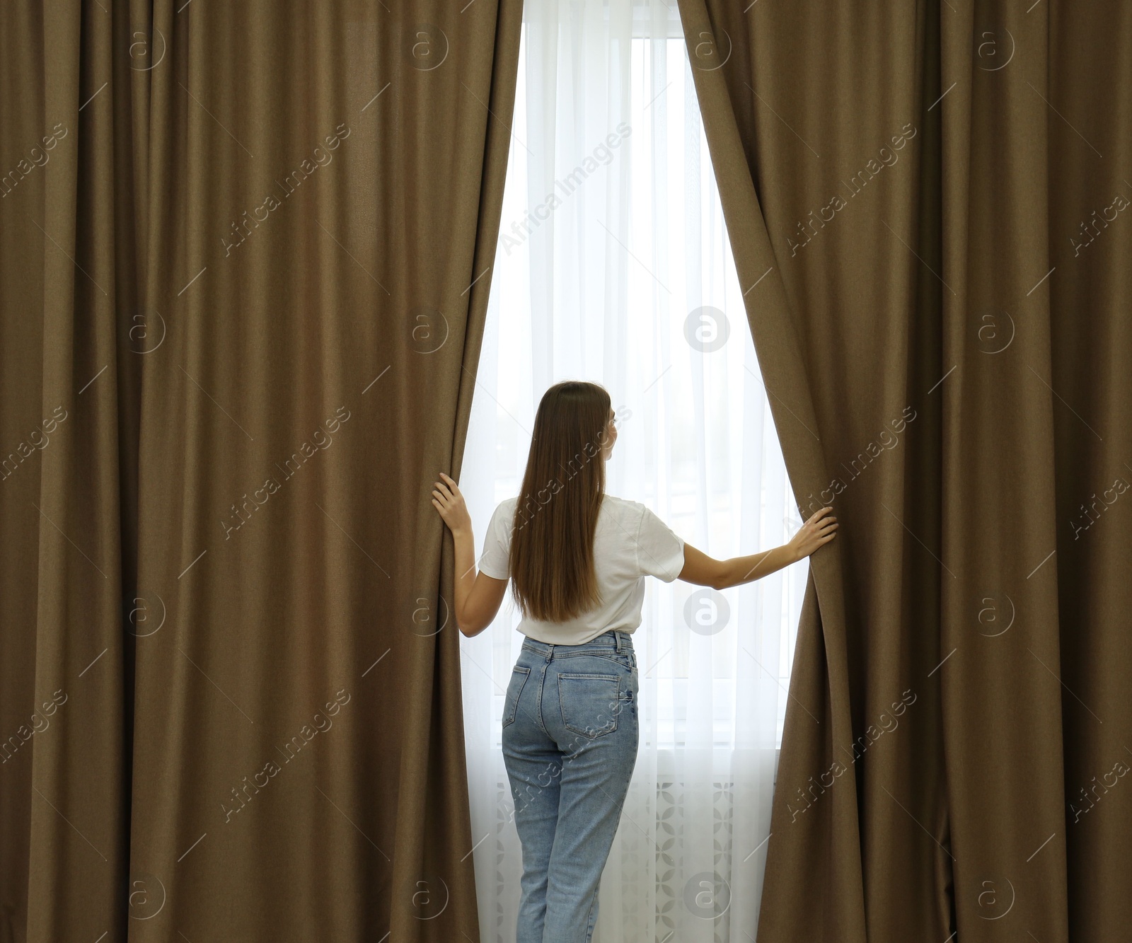 Woman opening elegant window curtains in room, back view Photo of Woman opening elegant window curtains in room, back view