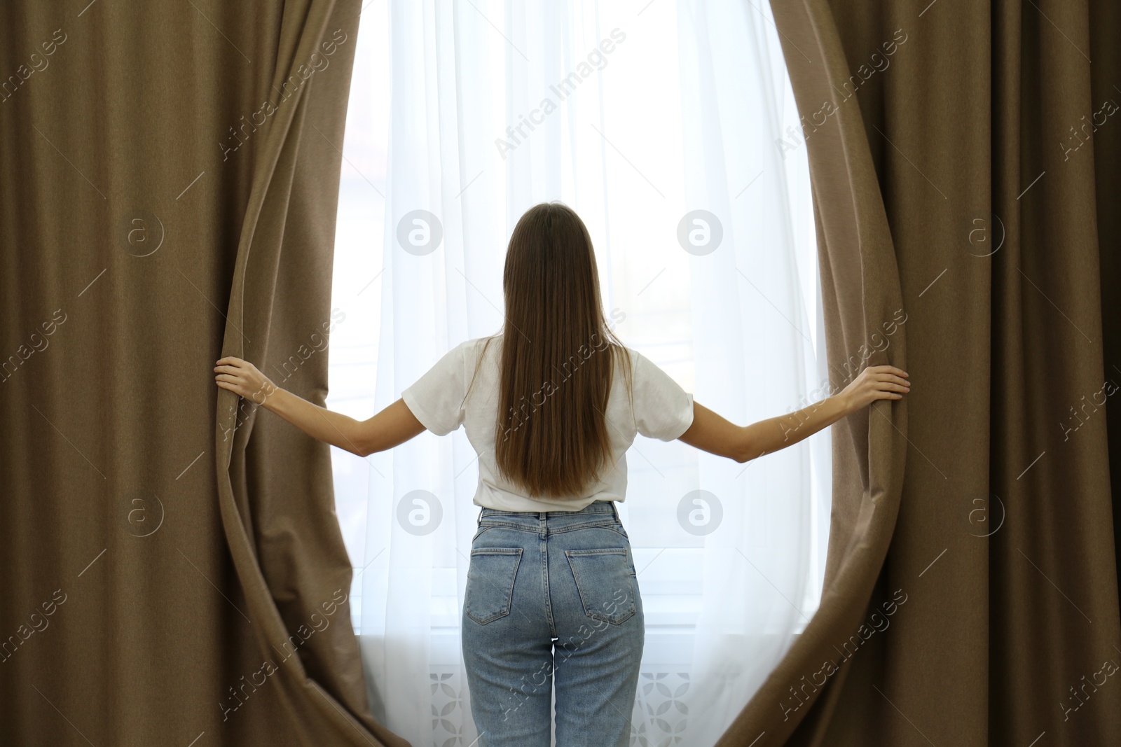 Woman opening elegant window curtains in room, back view Photo of Woman opening elegant window curtains in room, back view