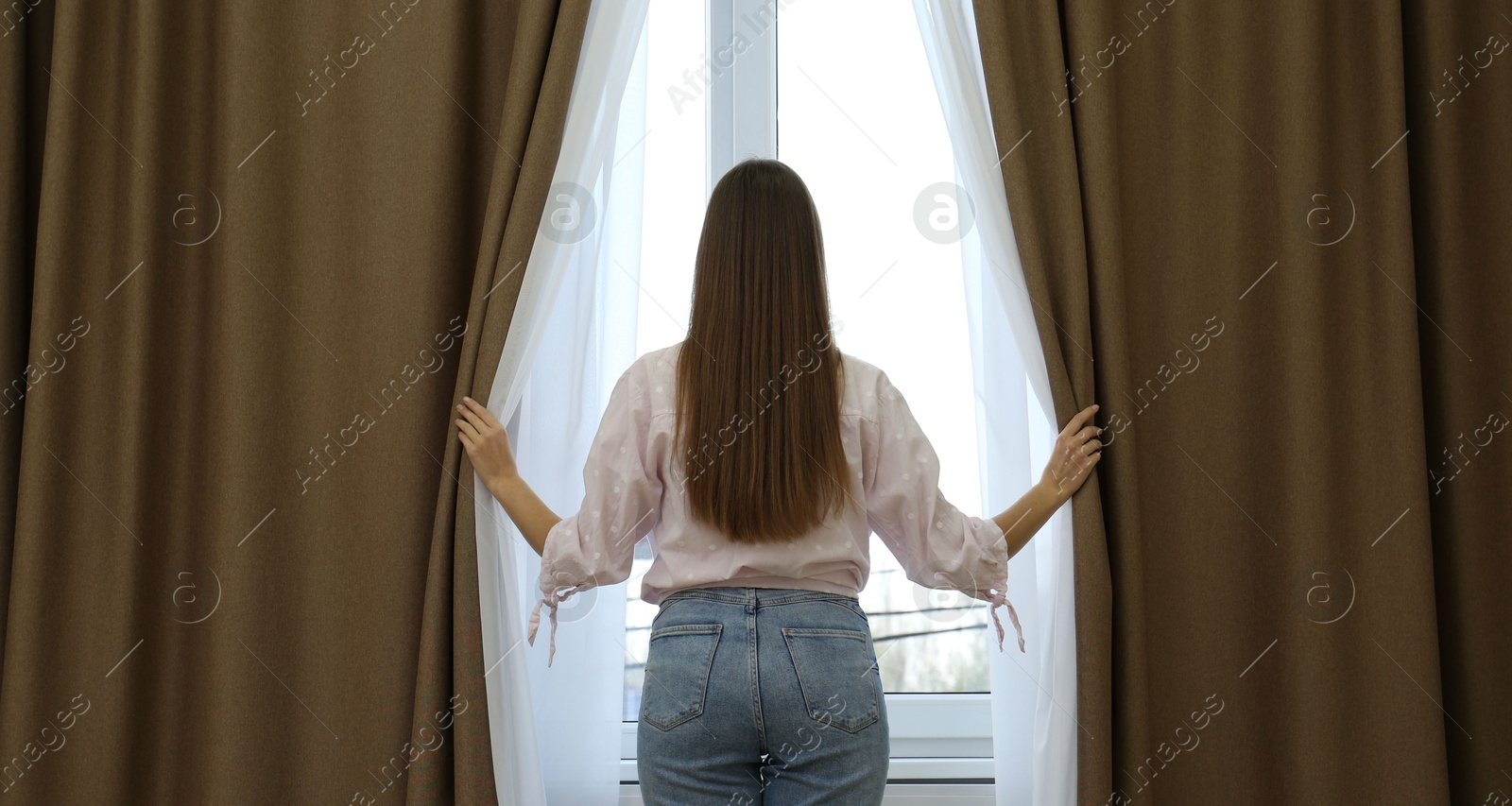 Woman opening elegant window curtains in room, back view Photo of Woman opening elegant window curtains in room, back view