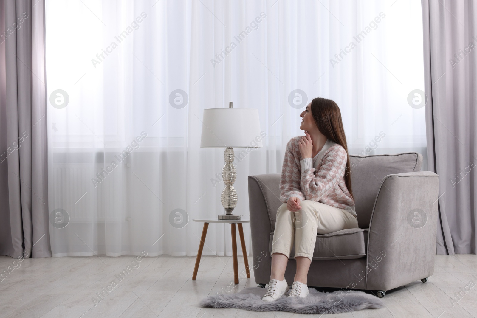 Woman sitting on armchair near lamp and window with stylish curtains at home. Space for text Photo of Woman sitting on armchair near lamp and window with stylish curtains at home. Space for text