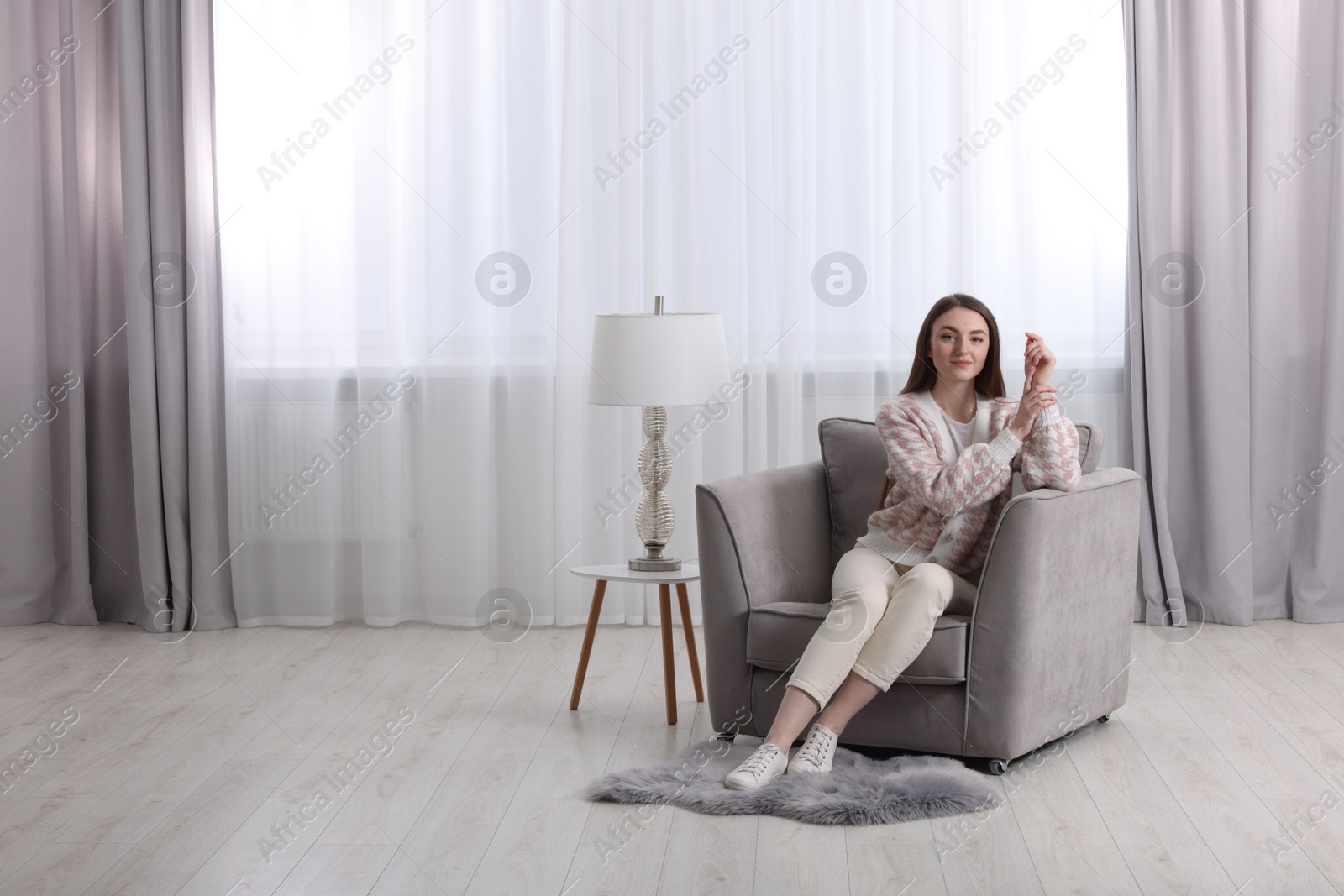 Woman sitting on armchair near lamp and window with stylish curtains at home. Space for text Photo of Woman sitting on armchair near lamp and window with stylish curtains at home. Space for text