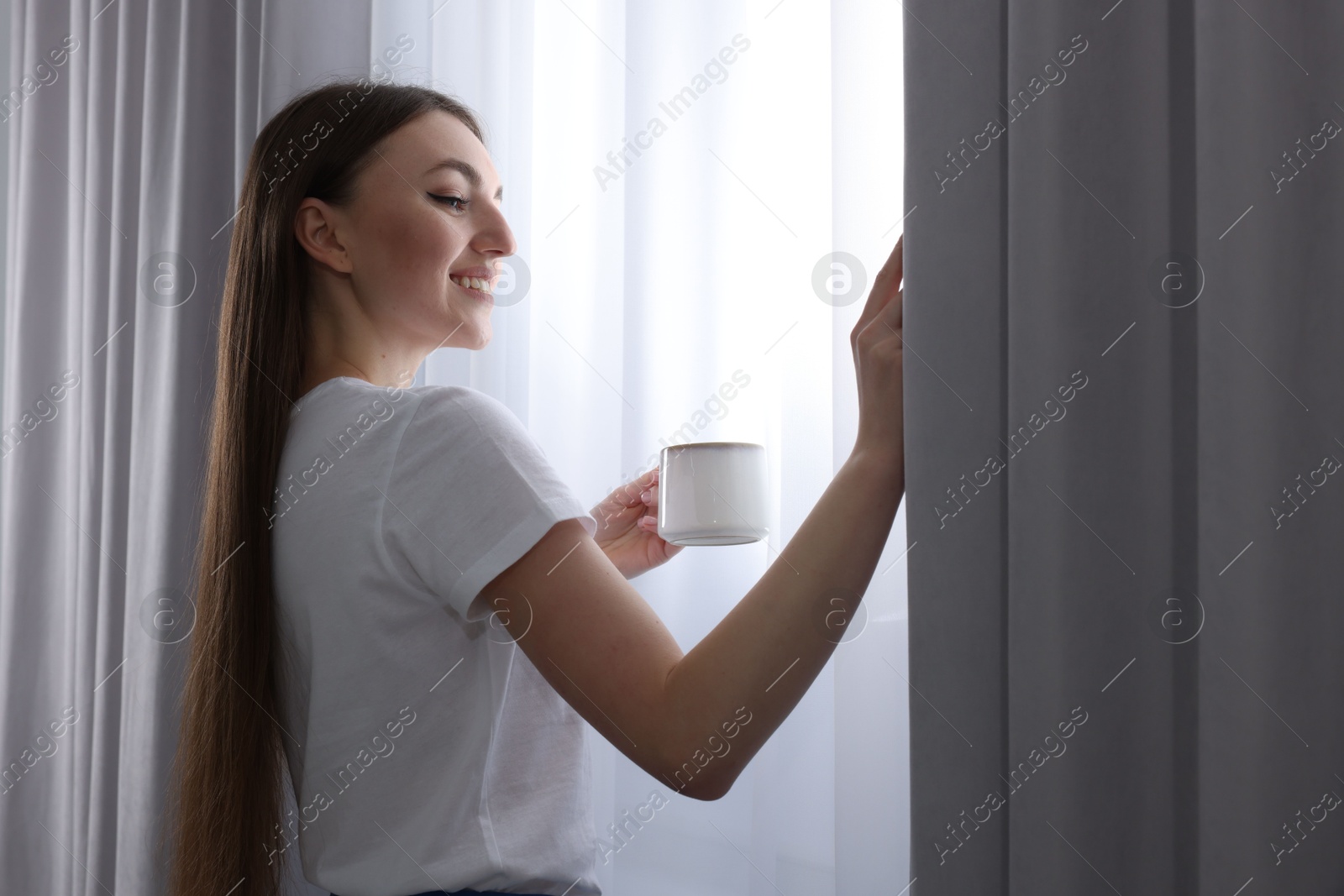 Woman with cup of hot drink opening stylish curtains at home Photo of Woman with cup of hot drink opening stylish curtains at home