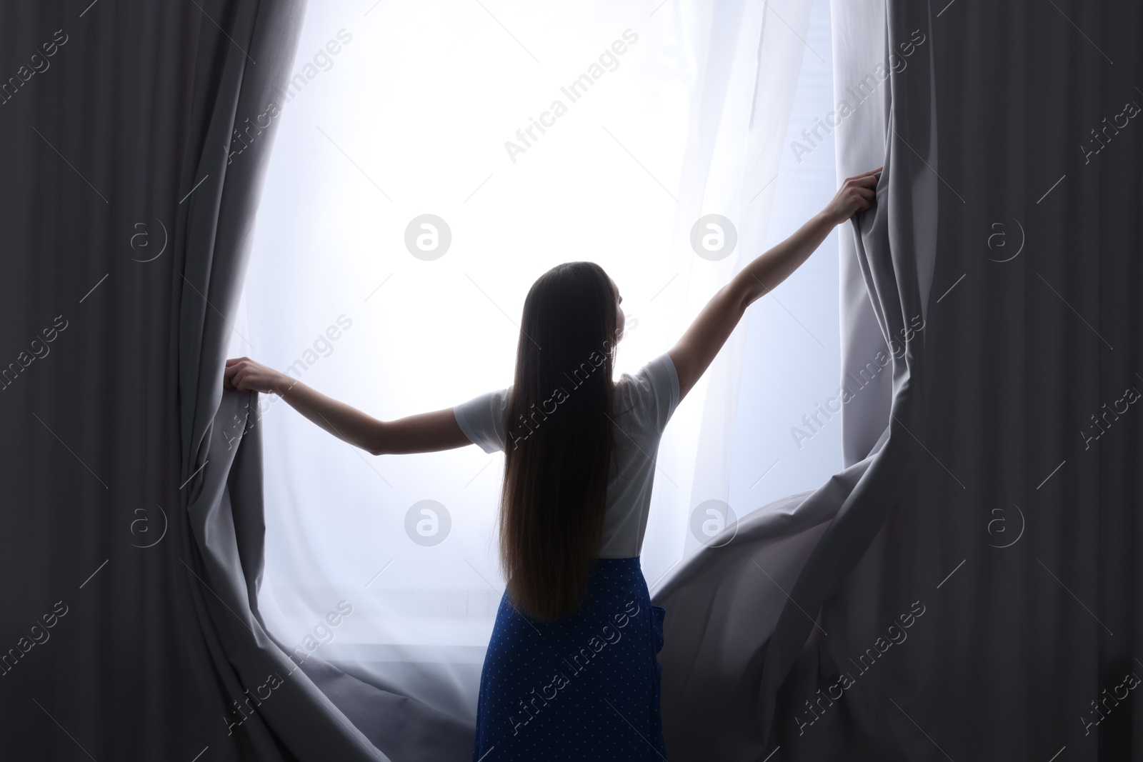 Woman opening stylish curtains at home, back view Photo of Woman opening stylish curtains at home, back view
