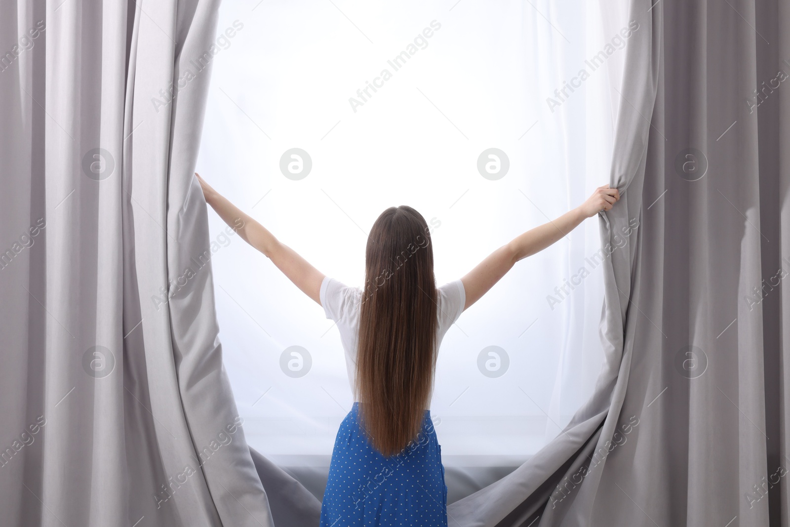 Woman opening stylish curtains at home, back view Photo of Woman opening stylish curtains at home, back view