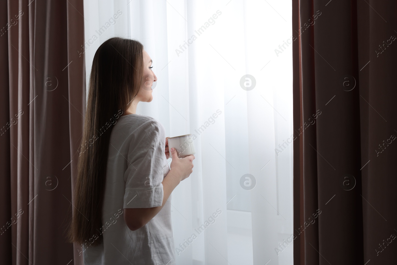 Woman holding cup of hot drink near window with stylish curtains at home. Space for text Photo of Woman holding cup of hot drink near window with stylish curtains at home. Space for text