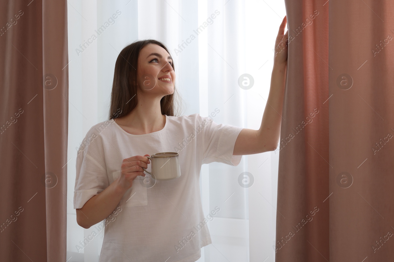 Woman with cup of hot drink opening stylish curtains at home Photo of Woman with cup of hot drink opening stylish curtains at home