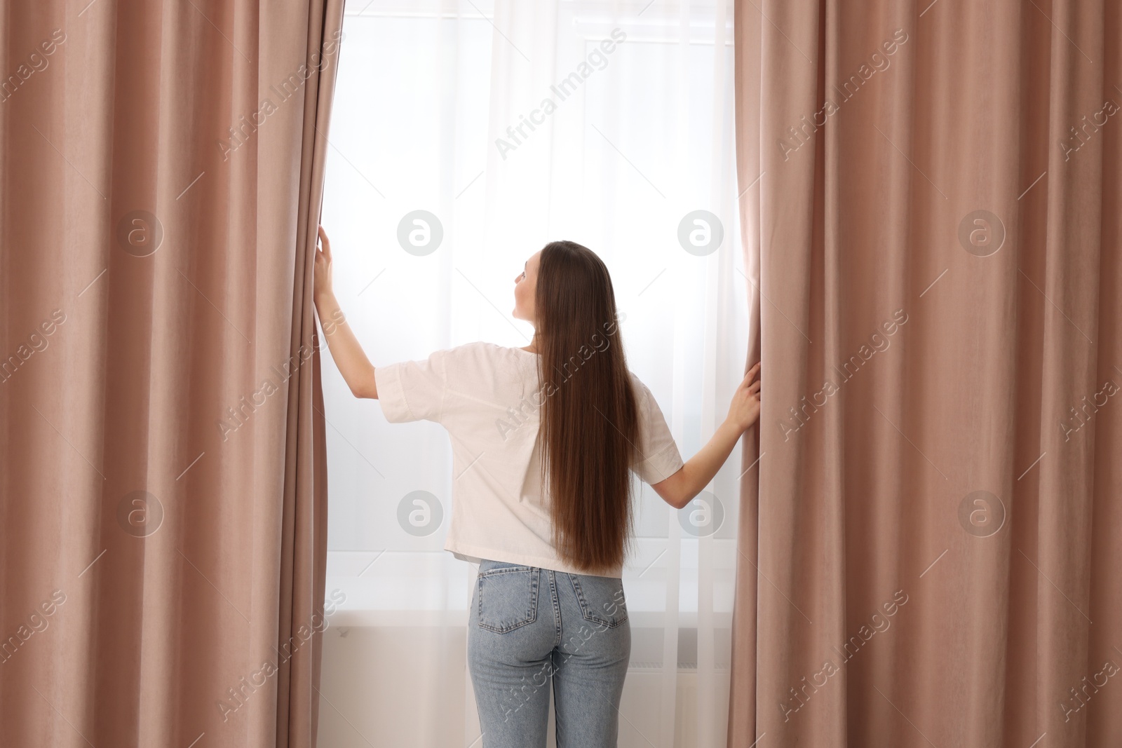 Woman opening stylish curtains at home, back view Photo of Woman opening stylish curtains at home, back view