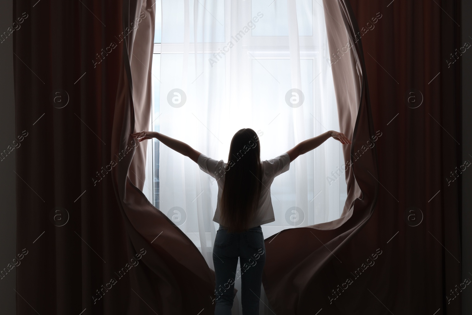 Woman opening stylish curtains at home, back view Photo of Woman opening stylish curtains at home, back view