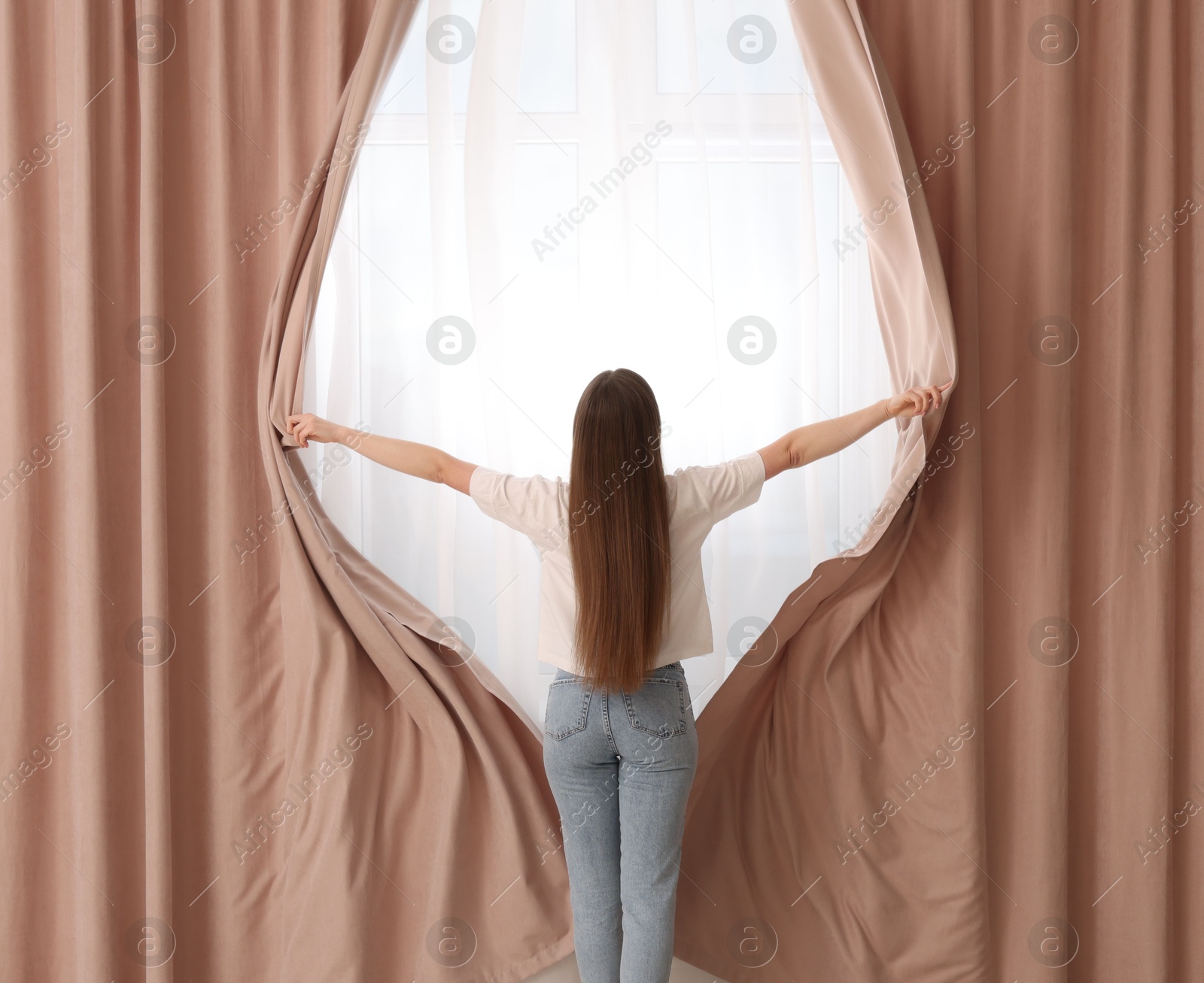 Woman opening stylish curtains at home, back view Photo of Woman opening stylish curtains at home, back view