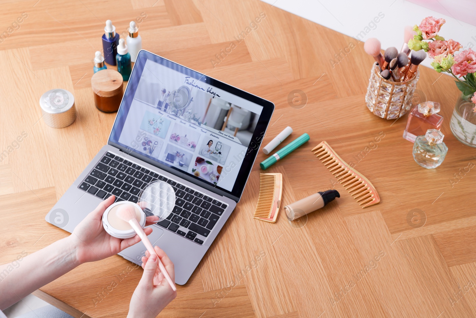 Blogger holding face powder and brush near laptop with fashion blog web site at wooden table indoors, above view Photo of Blogger holding face powder and brush near laptop with fashion blog web site at wooden table indoors, above view