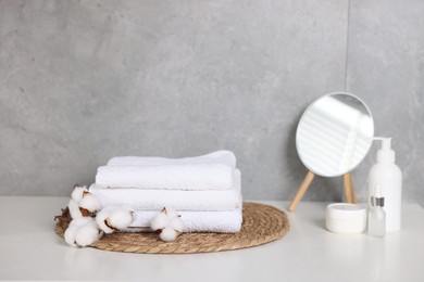 Photo of Stack of clean towels, cotton flowers, cosmetic products and mirror on white table indoors