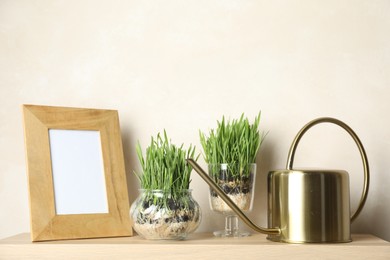 Photo of Wheat grass in transparent pots, frame and watering can on wooden table near light wall. Space for text