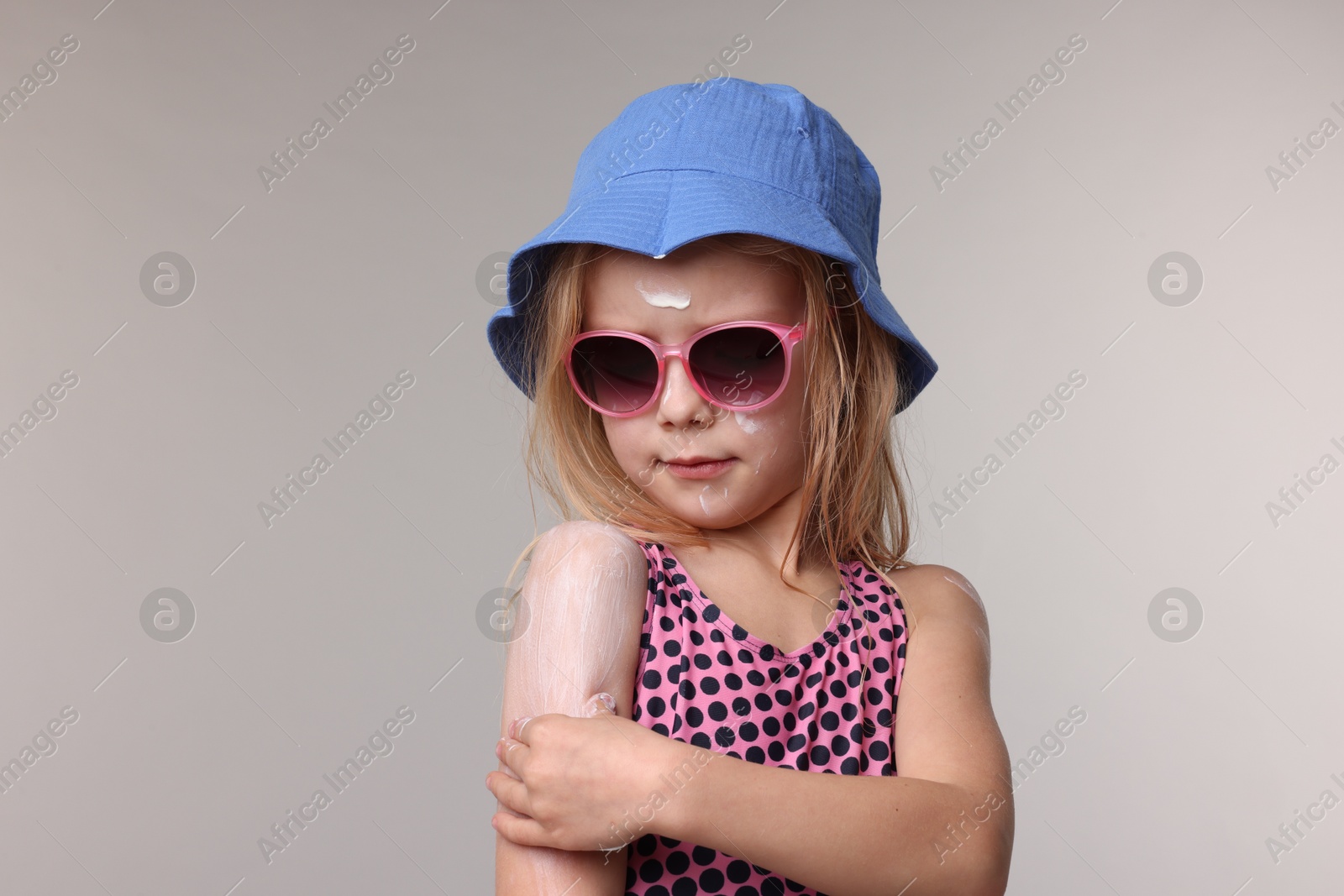 Photo of Cute little girl applying sun protection product onto her shoulder on grey background