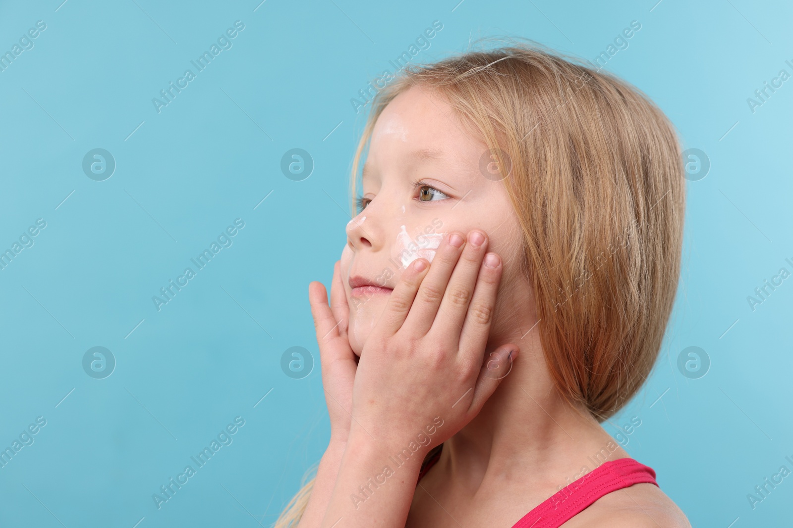 Photo of Cute little girl applying sun protection cream onto her face against light blue background, closeup. Space for text