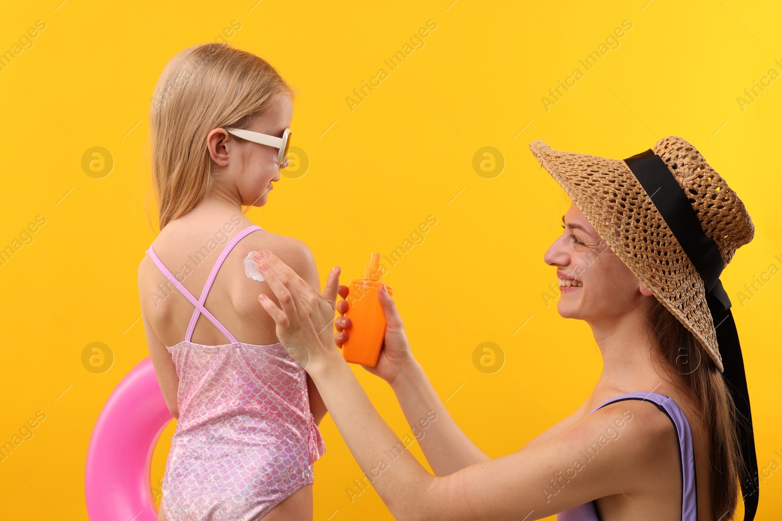 Smiling mother applying sun protection product onto her daughter's skin on yellow background, back view Photo of Smiling mother applying sun protection product onto her daughter's skin on yellow background, back view