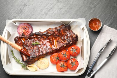 Photo of Delicious spare ribs, baked onion, sun dried tomatoes and sauce on grey textured table, flat lay