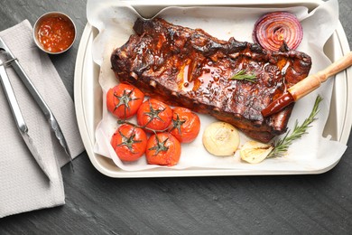 Photo of Delicious spare ribs, baked onion, sun dried tomatoes and sauce on grey textured table, flat lay