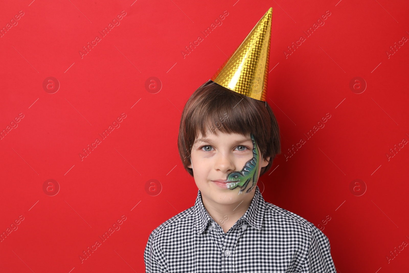 Cute boy with dinosaur face painting and party hat on red background, space for text Photo of Cute boy with dinosaur face painting and party hat on red background, space for text