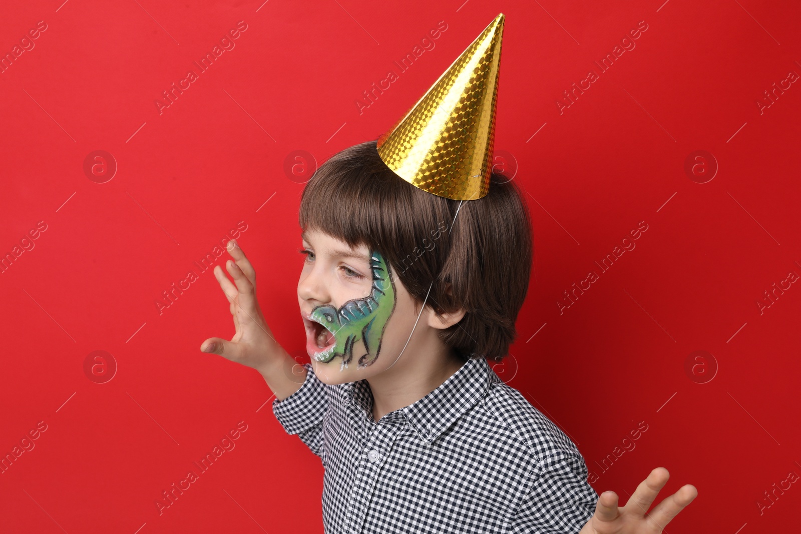 Cute boy with dinosaur face painting and party hat posing on red background Photo of Cute boy with dinosaur face painting and party hat posing on red background