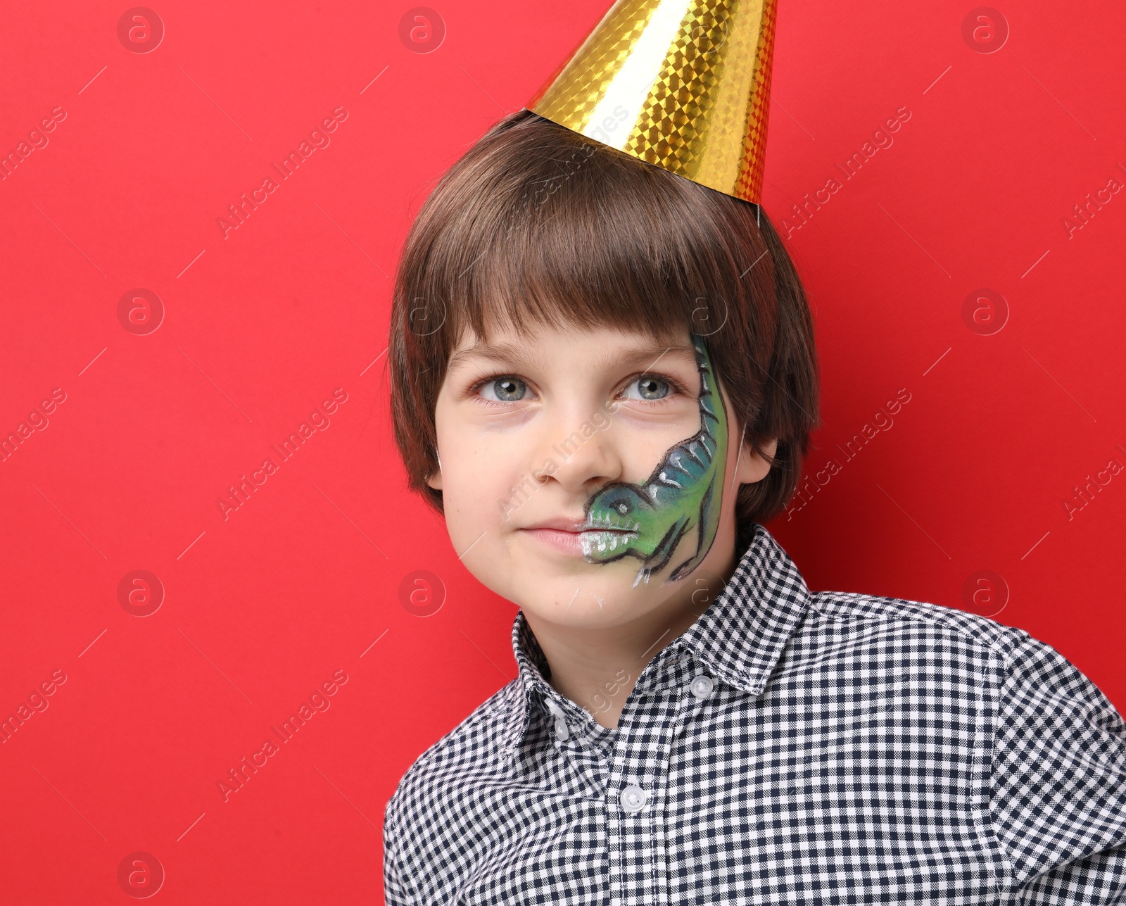 Cute boy with dinosaur face painting and party hat on red background Photo of Cute boy with dinosaur face painting and party hat on red background
