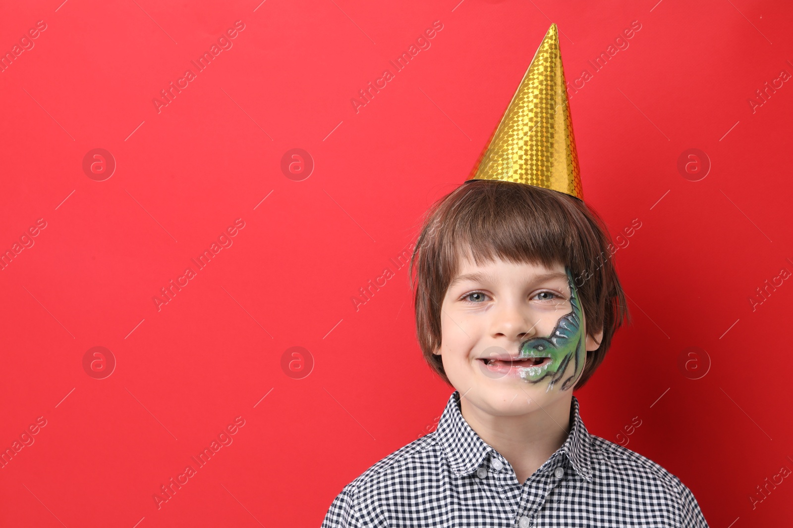 Cute boy with dinosaur face painting and party hat on red background, space for text Photo of Cute boy with dinosaur face painting and party hat on red background, space for text