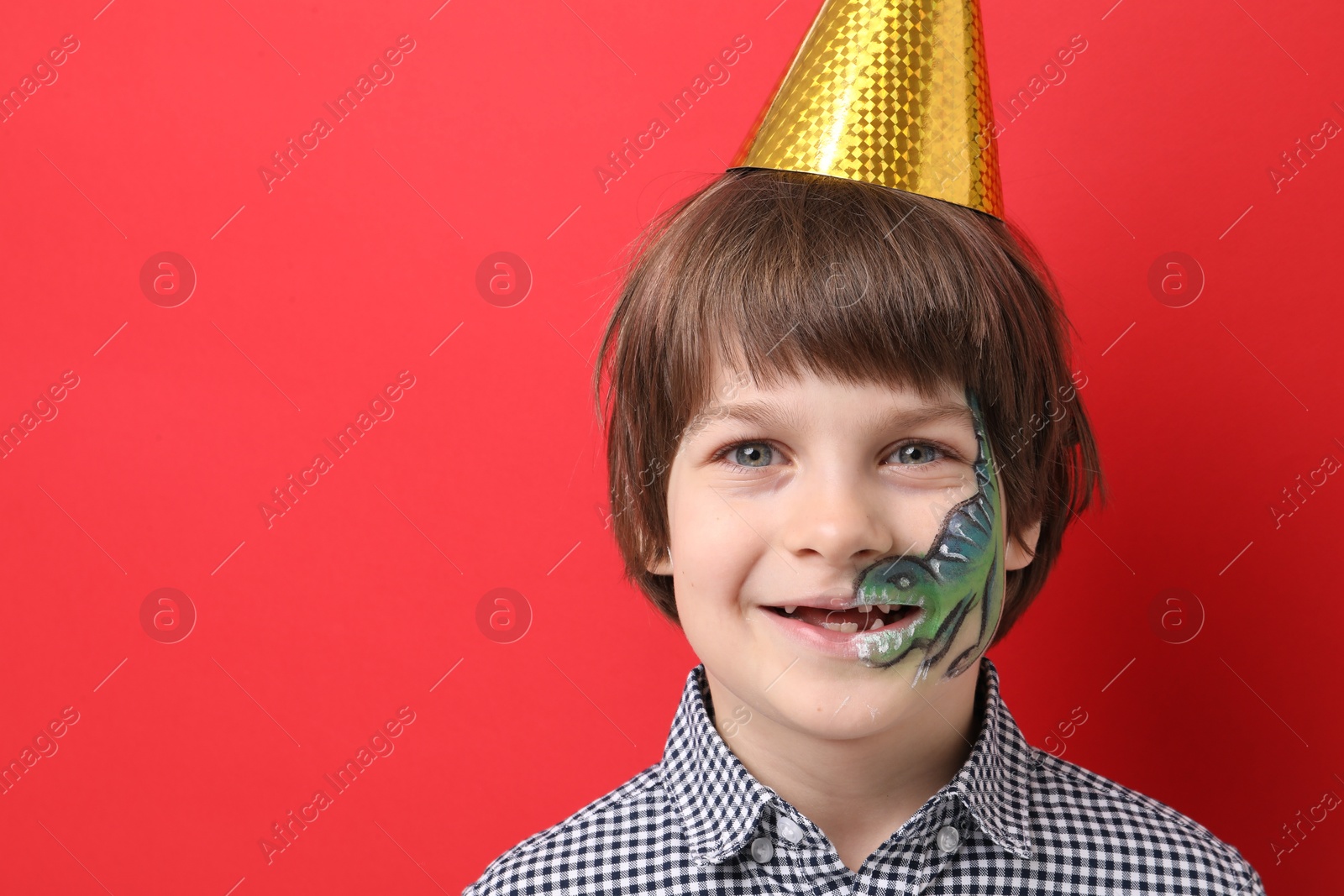Cute boy with dinosaur face painting and party hat on red background, space for text Photo of Cute boy with dinosaur face painting and party hat on red background, space for text