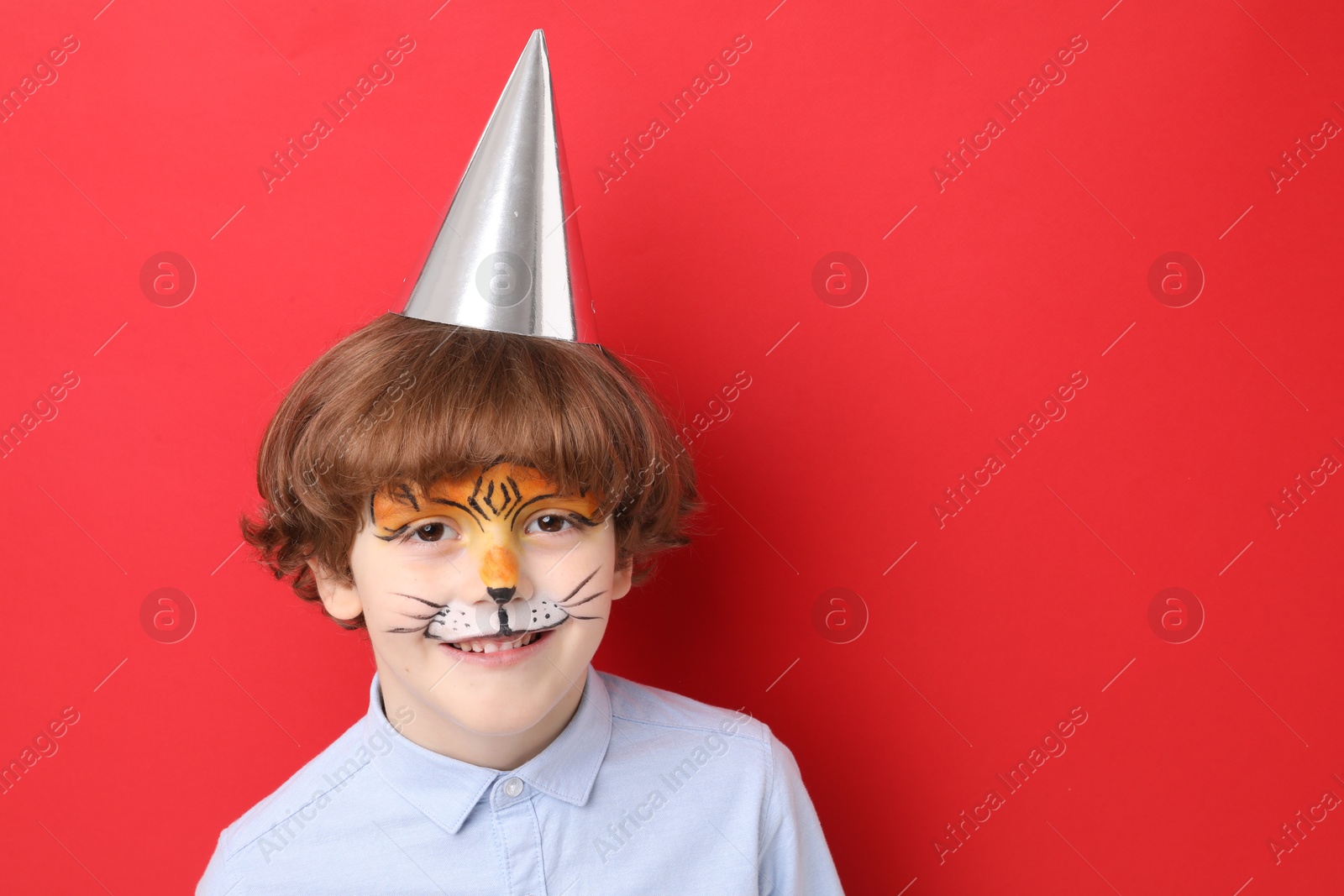 Cute boy with tiger face painting and party hat on red background, space for text Photo of Cute boy with tiger face painting and party hat on red background, space for text