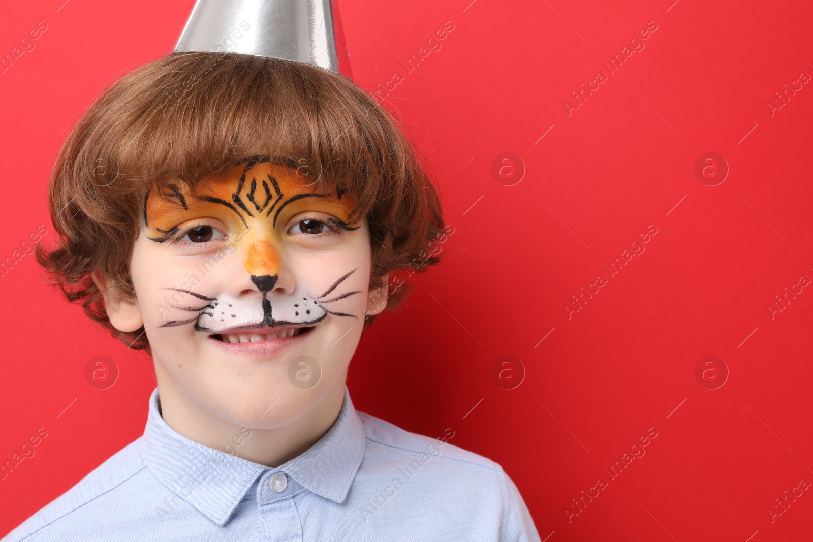 Cute boy with tiger face painting and party hat on red background, space for text Photo of Cute boy with tiger face painting and party hat on red background, space for text