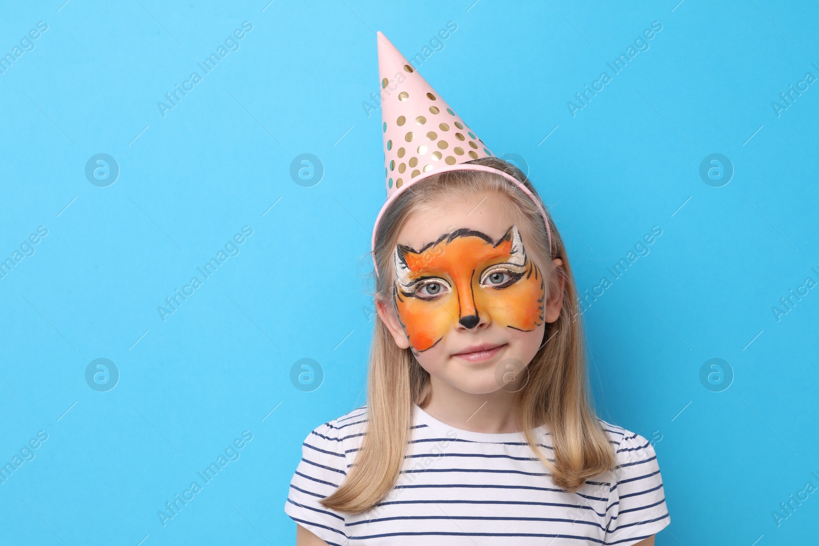 Cute girl with fox face painting and party hat on light blue background, space for text Photo of Cute girl with fox face painting and party hat on light blue background, space for text