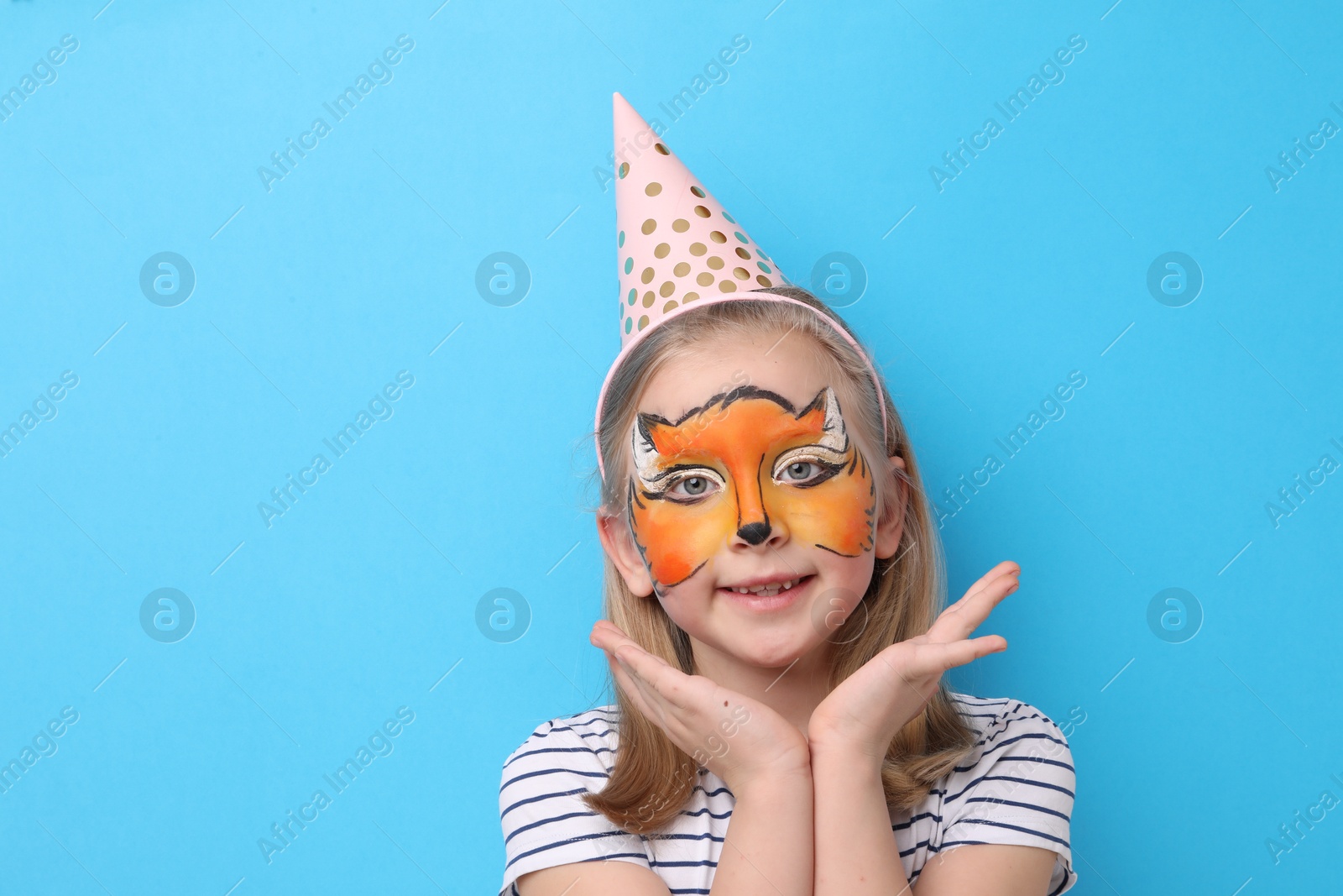 Cute girl with fox face painting and party hat on light blue background, space for text Photo of Cute girl with fox face painting and party hat on light blue background, space for text