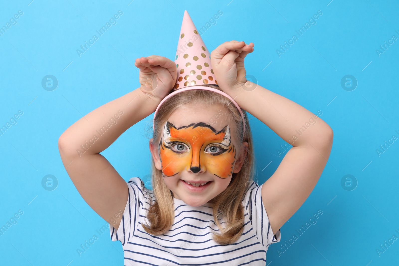 Cute girl with fox face painting and party hat posing on light blue background Photo of Cute girl with fox face painting and party hat posing on light blue background