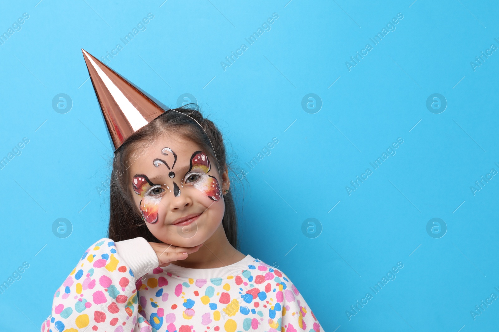 Cute girl with butterfly face painting and party hat on light blue background, space for text Photo of Cute girl with butterfly face painting and party hat on light blue background, space for text