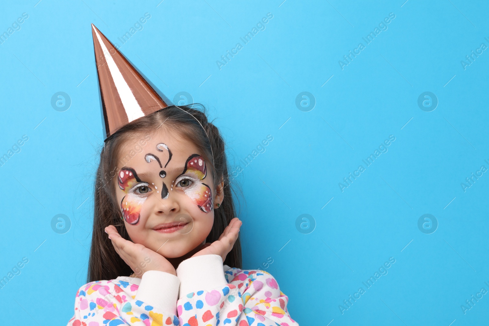 Cute girl with butterfly face painting and party hat on light blue background, space for text Photo of Cute girl with butterfly face painting and party hat on light blue background, space for text