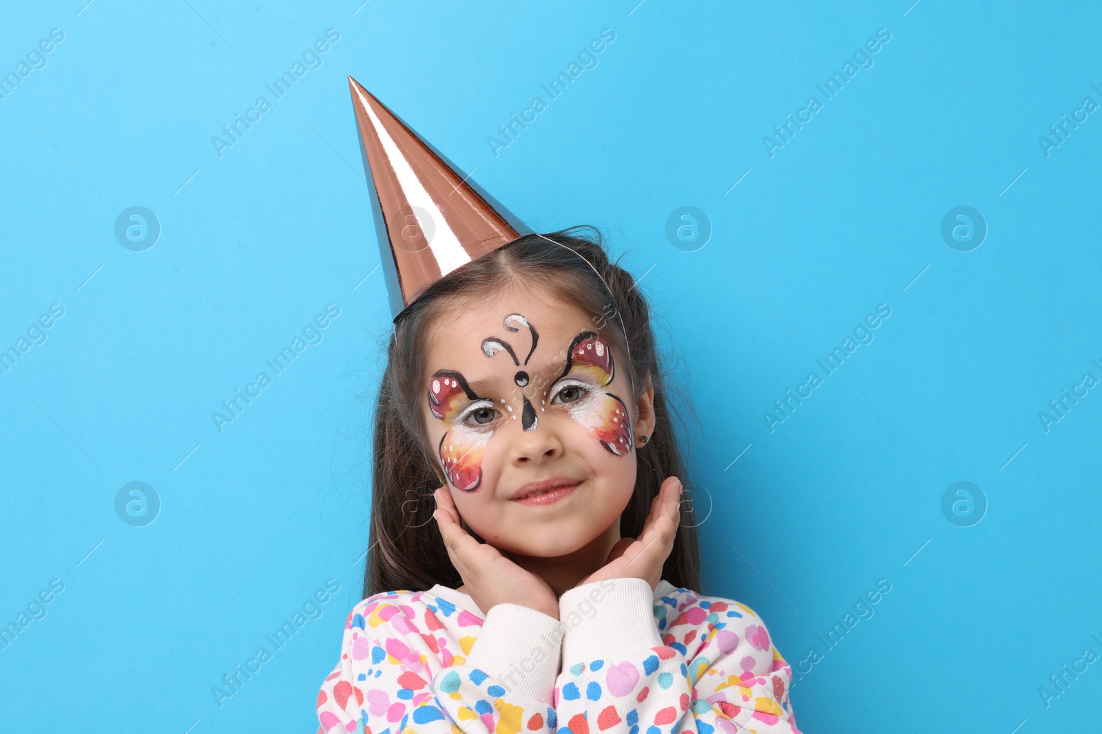 Cute girl with butterfly face painting and party hat on light blue background Photo of Cute girl with butterfly face painting and party hat on light blue background