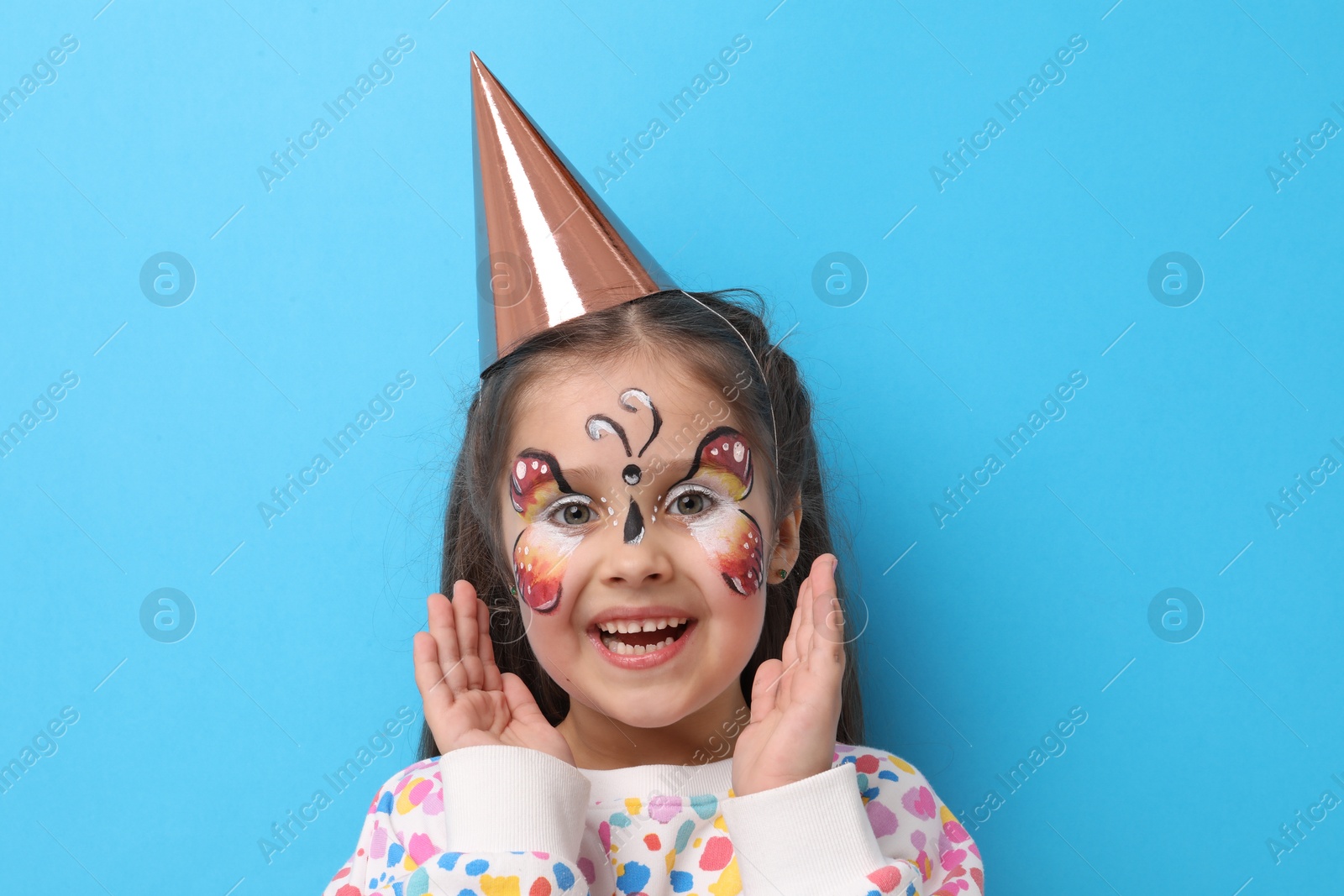 Cute girl with butterfly face painting and party hat on light blue background Photo of Cute girl with butterfly face painting and party hat on light blue background