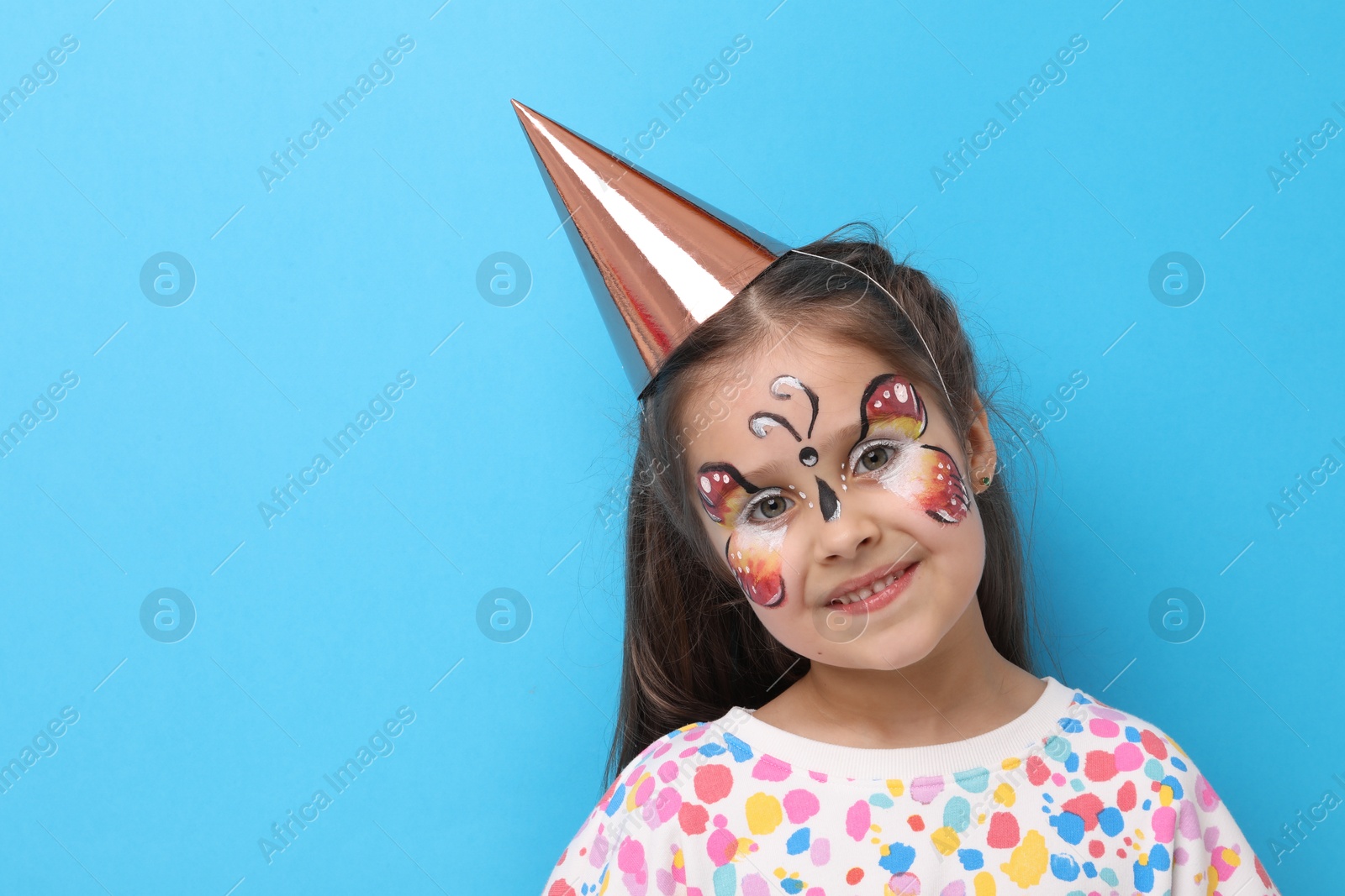 Cute girl with butterfly face painting and party hat on light blue background, space for text Photo of Cute girl with butterfly face painting and party hat on light blue background, space for text