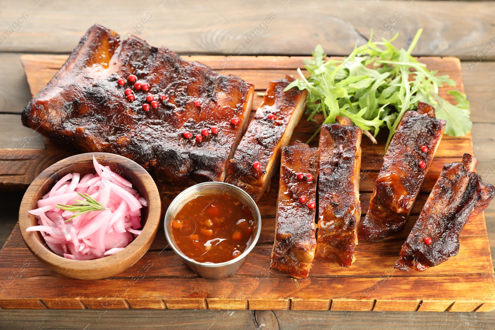 Delicious spare ribs, onion, sauce and arugula on wooden table Photo of Delicious spare ribs, onion, sauce and arugula on wooden table