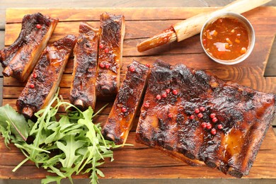 Photo of Delicious spare ribs, sauce, arugula and brush on wooden table, top view