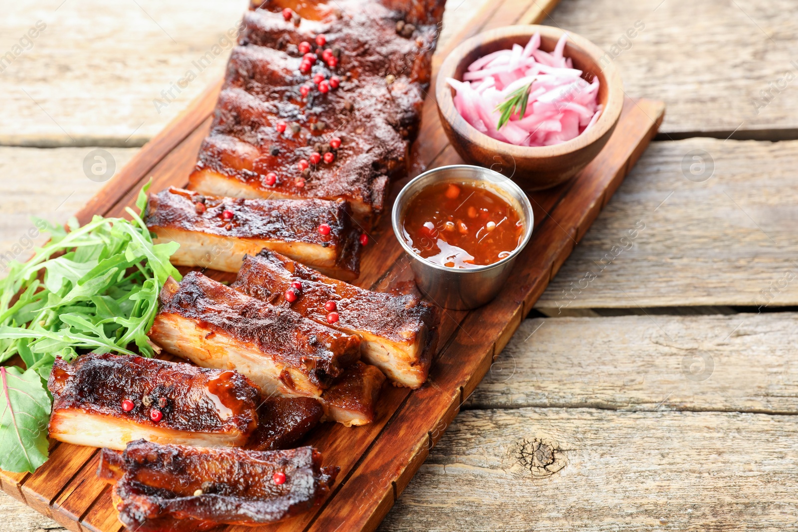Delicious spare ribs, onion, sauce and arugula on wooden table Photo of Delicious spare ribs, onion, sauce and arugula on wooden table