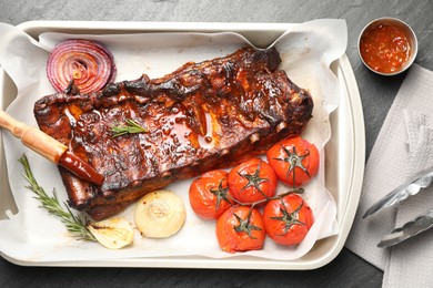 Photo of Delicious spare ribs, rosemary, baked onion, sauce and sun dried tomatoes on grey textured table, flat lay