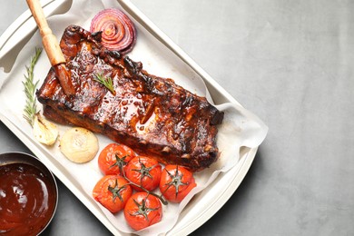Photo of Delicious spare ribs, rosemary, baked onion, sauce and sun dried tomatoes on grey table, flat lay. Space for text